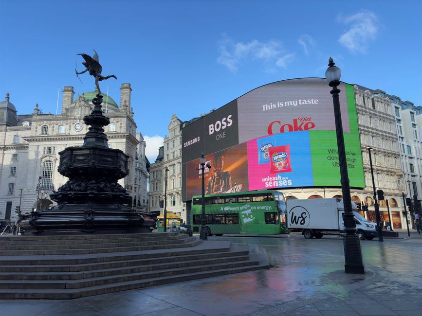 A daytime view of a busy London square featuring the Shaftesbury Memorial Fountain with the statue of Eros on top, situated on a circular platform with steps leading up. Behind the fountain, a large c