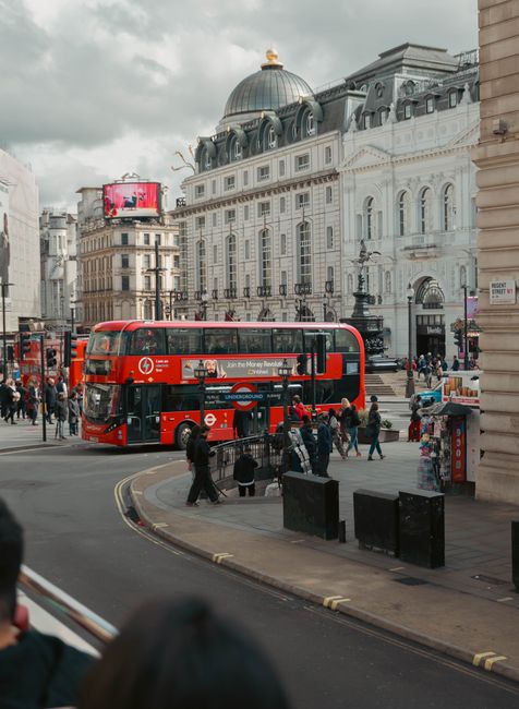 A daytime view of a busy London square featuring the Shaftesbury Memorial Fountain with the statue of Eros on top, situated on a circular platform with steps leading up. Behind the fountain, a large c