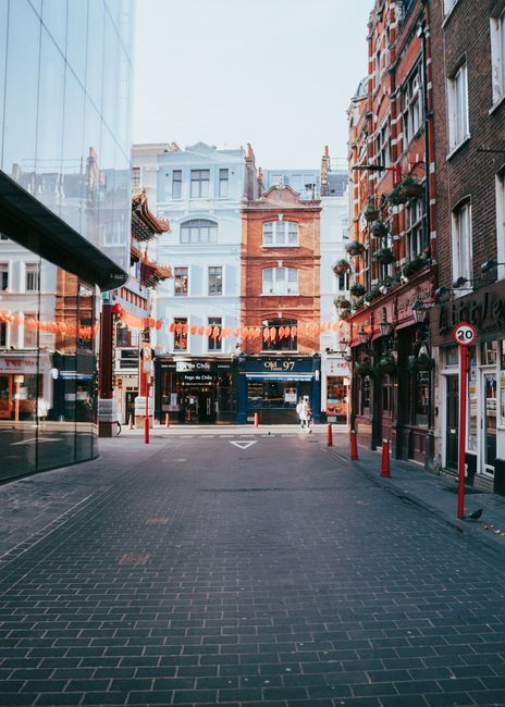 Night-time view of a narrow cobblestone street in Soho, London, decorated with string lights hanging across from side to side between brick buildings. The buildings feature large windows, some with wi