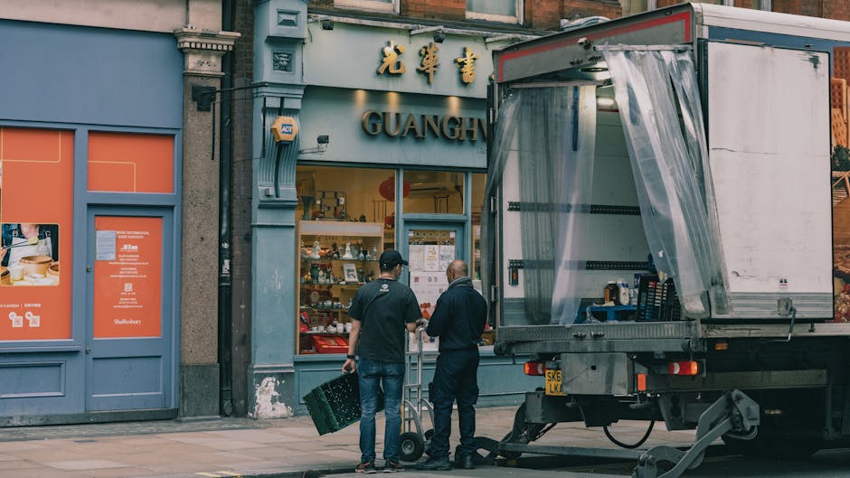 Two men dressed in casual work clothing are engaged in a home relocation process outside a storefront. One man, wearing a black cap and jeans, is carrying a plastic crate, while the other, with a bald